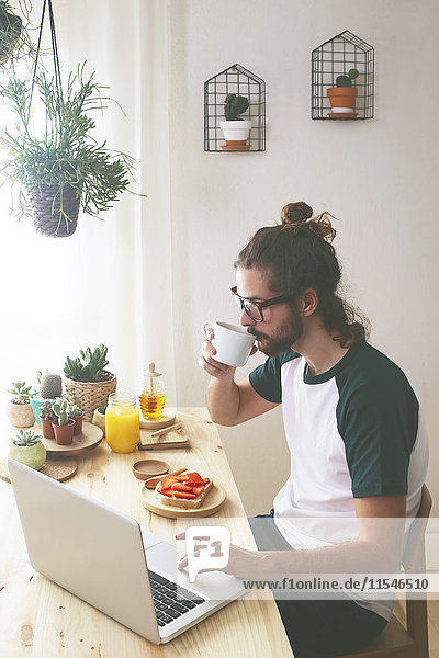 Junger Mann mit Laptop beim Frühstücken