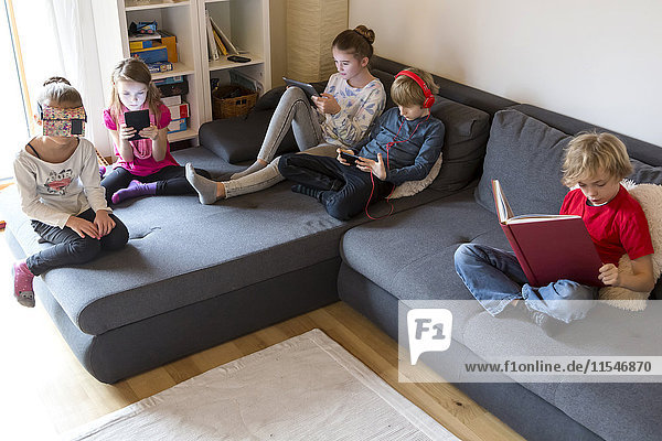 Vier Kinder auf einer Couch mit verschiedenen digitalen Geräten  während ein Junge ein Buch liest.