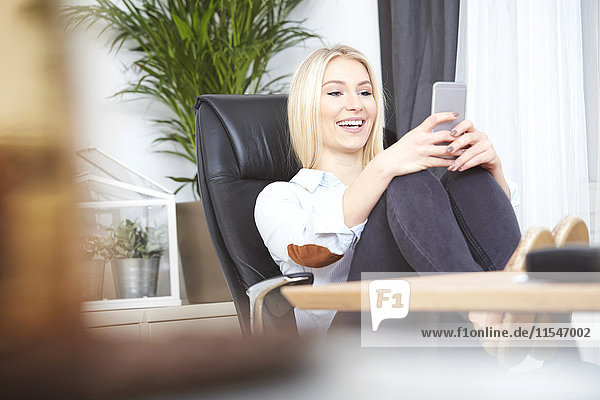 Porträt einer lächelnden blonden Frau  die an ihrem Schreibtisch sitzt und mit erhobenen Füßen einen Selfie mit dem Smartphone nimmt.