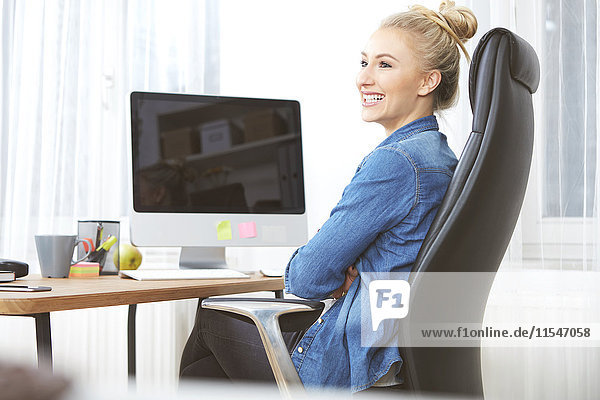 Lächelnde blonde Frau sitzt am Schreibtisch in ihrem Heimbüro.