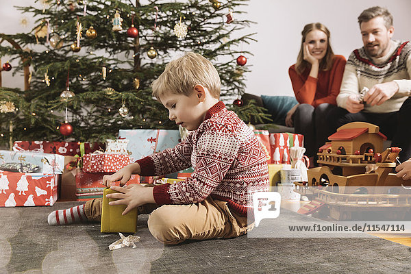 Kleiner Junge eröffnet Weihnachtsgeschenk mit Eltern im Hintergrund