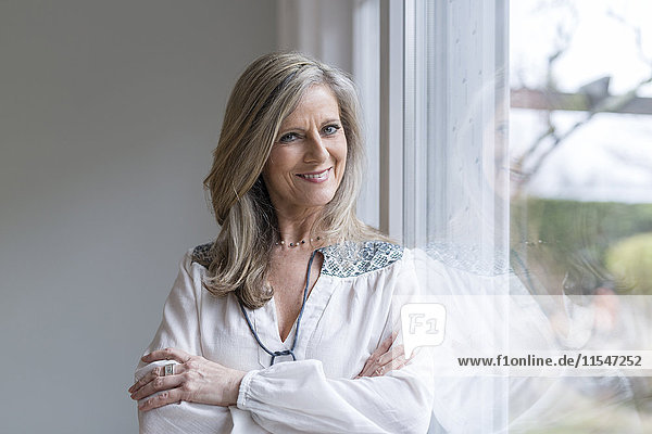 Portrait of smiling blond woman with arms crossed leaning against window