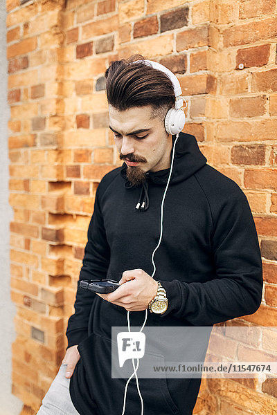 Portrait of young man leaning against brick wall listening music with headphones
