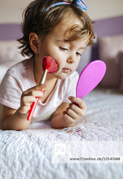 Porträt eines kleinen Mädchens auf dem Bett liegend mit Handspiegel und Kosmetikpinsel