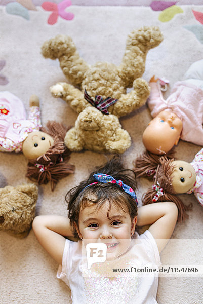 Porträt eines lächelnden kleinen Mädchens auf dem Boden liegend mit Teddys und Puppen um sie herum.