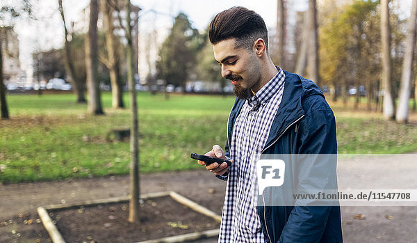 Lächelnder junger Mann mit Blick auf sein Smartphone im Park