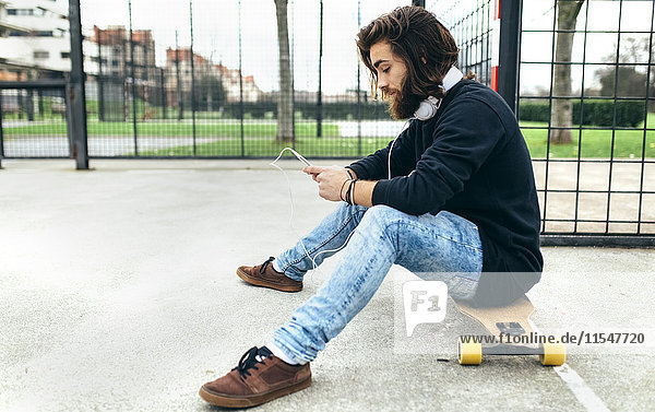 Junge bärtige Skateboarderin mit Smartphone und Kopfhörer