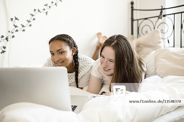 Two teenage girls lying on bed looking at laptop