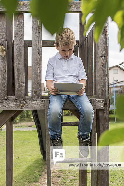 Kleiner Junge sitzt auf einem Spielplatz und schaut auf ein digitales Tablett.