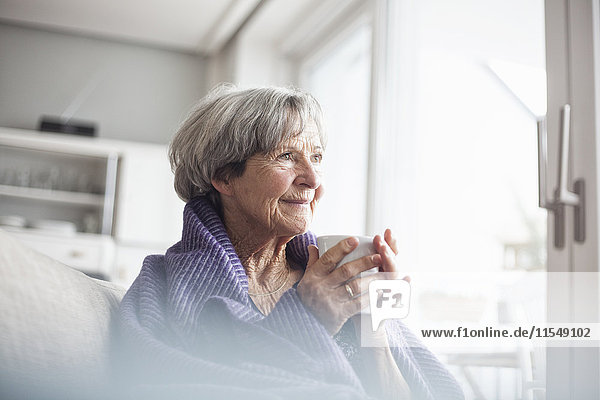 Porträt einer fröhlichen Seniorin auf der Couch zu Hause bei einer Tasse Kaffee