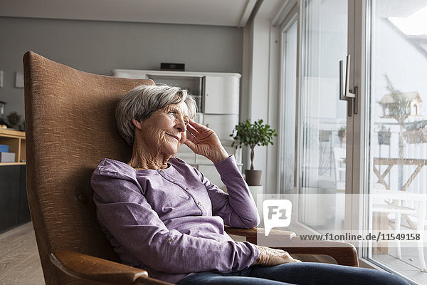 Porträt einer älteren Frau  die zu Hause auf einem Sessel sitzt und durchs Fenster schaut.
