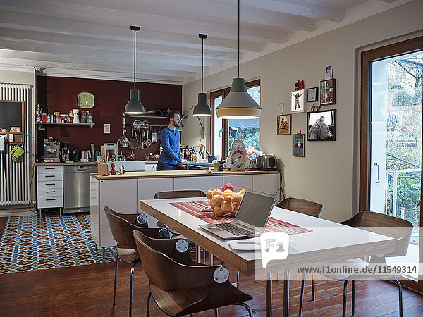 Laptop on dining table in open plan kitchen with man in background