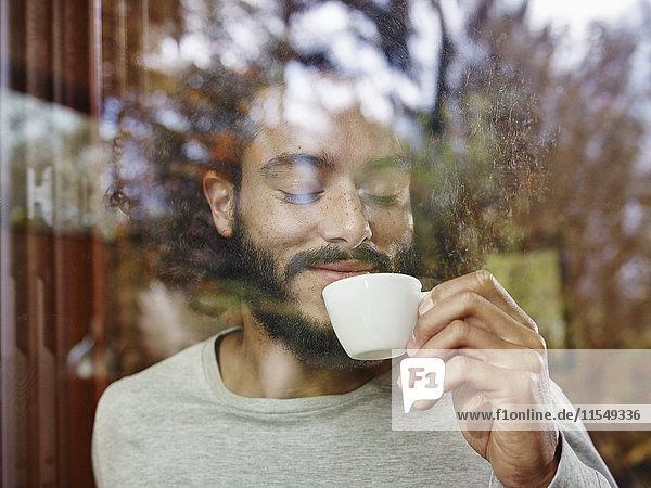 Smiling young man enjoying cup of coffee behind windowpane