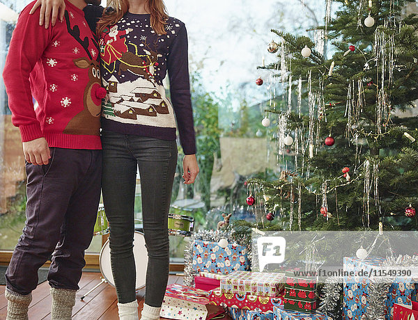 Ein Paar steht vor dem Weihnachtsbaum und trägt Weihnachtspullover.