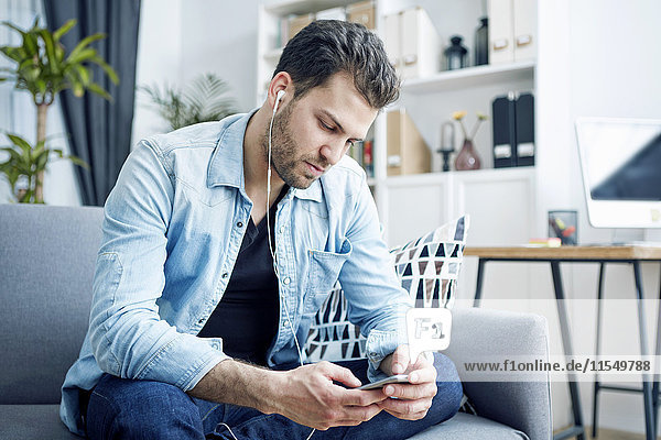 Junger Mann zu Hause mit Ohrhörern beim Blick auf das Smartphone