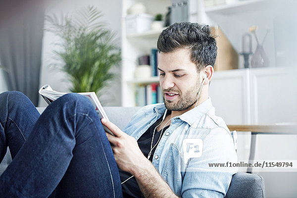 Young man at home with earbuds reading magazine