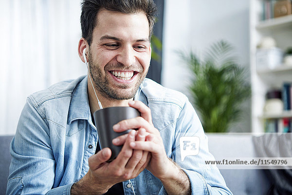 Happy young man at home with earbuds holding cup