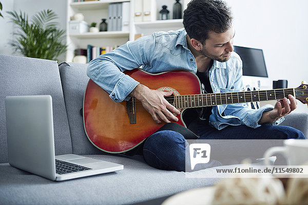 Junger Mann zu Hause sitzt auf der Couch und spielt Gitarre neben dem Laptop.