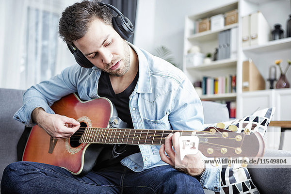 Young man at home sitting on couch wearing headphones and playing guitar