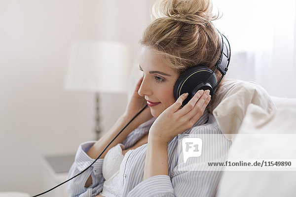 Porträt einer lächelnden blonden Frau  die auf dem Bett sitzt und Musik mit Kopfhörern hört.