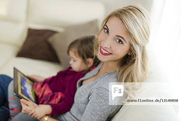 Porträt einer lächelnden Frau  die mit ihrer kleinen Tochter auf der Couch im Wohnzimmer sitzt.
