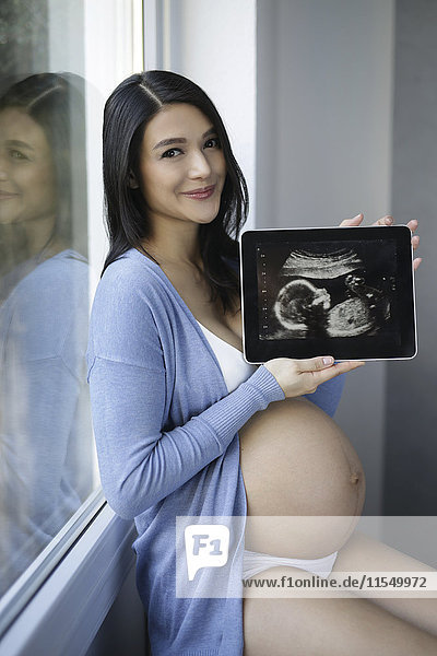 Lächelnde schwangere Frau sitzt am Fenster und zeigt Ultraschall auf digitalem Tablett.