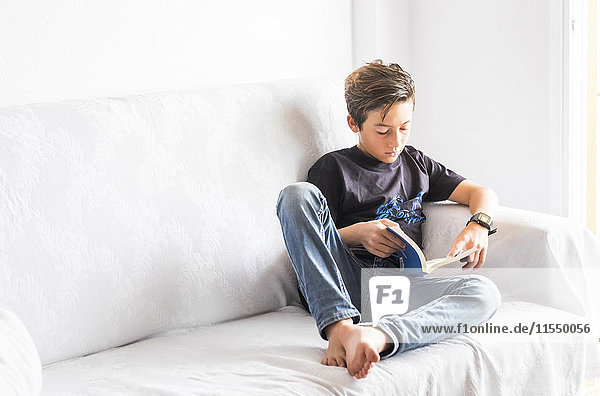 Junge sitzt auf einer weißen Couch und liest ein Buch.