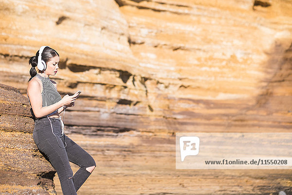 Spanien  Teneriffa  junge Frau mit Kopfhörer  die vor einer Felswand steht und ihr Smartphone ansieht.