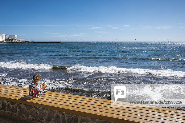 Spanien  Teneriffa  Frau auf einer Bank vor dem Meer sitzend mit ihrem Smartphone