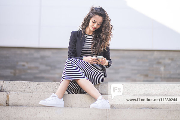 Porträt eines jungen Mädchens  das auf einer Treppe sitzt und auf sein Smartphone schaut.
