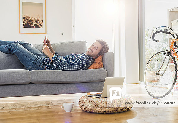 Junger Mann mit digitalem Tablett zu Hause auf der Couch liegend