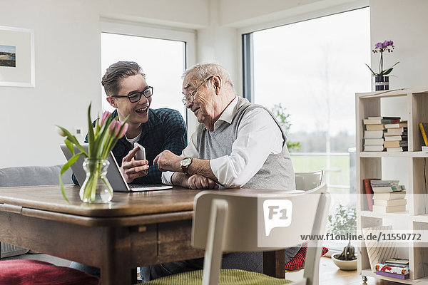 Senior Mann und sein Enkel sitzen am Tisch im Wohnzimmer mit Laptop und Smartphone