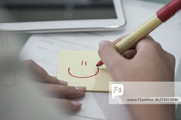 Nahaufnahme einer Frau am Schreibtisch  die ein Smiley auf einer Haftnotiz zeichnet.