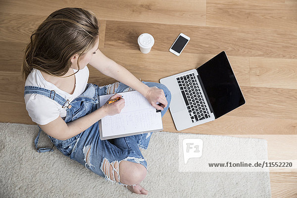Junge Frau sitzt auf dem Boden mit Laptop und Klemmbrett und macht sich Notizen.