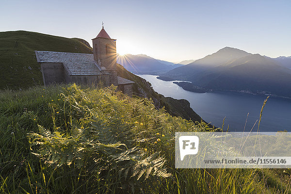 Sunbeam on Church of San Bernardo lights up the landscape around the blue water of Lake Como at dawn  Musso  Lombardy  Italy  Europe