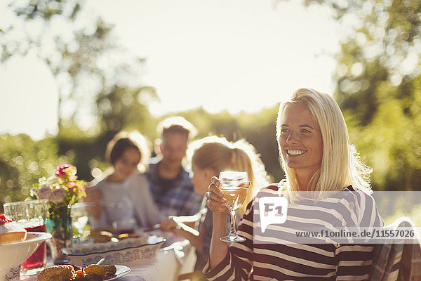 Lächelnde Frau trinkt Wein am sonnigen Gartenpartytisch