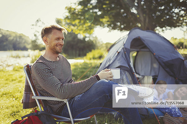 Lächelnder Vater trinkt Kaffee vor dem Zelt auf dem Campingplatz