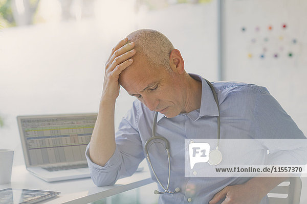 Müder  gestresster männlicher Arzt mit dem Kopf in den Händen in einer Arztpraxis