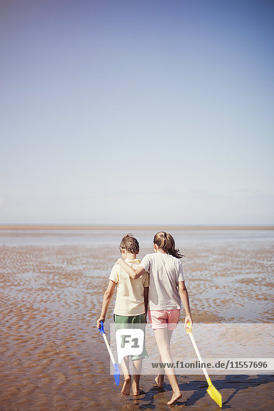 Bruder und Schwester mit Schaufeln  die sich umarmen und im nassen Sand am sonnigen Sommerstrand unter blauem Himmel laufen