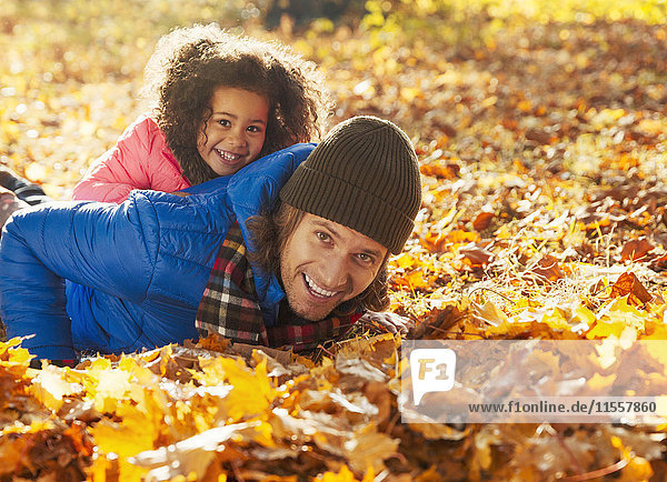 Portrait verspielte Tochter auf Vater liegend im Herbstlaub im sonnigen Park
