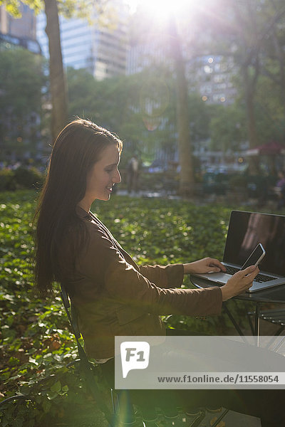 USA  New York  Manhattan  lächelnde junge Geschäftsfrau am Tisch im Park mit Blick auf das Handy