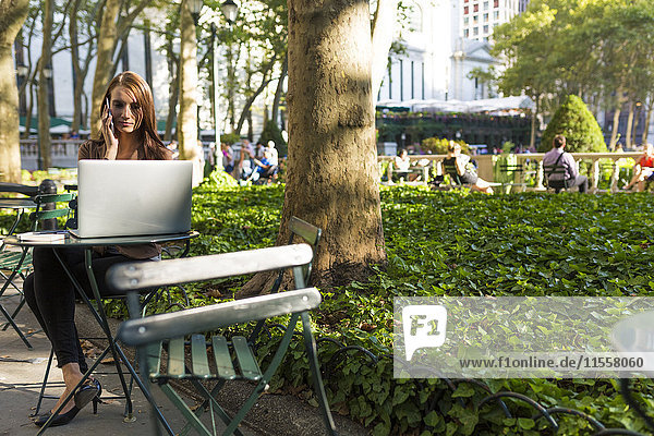 USA  New York  Manhattan  junge Geschäftsfrau am Telefon am Tisch im Park mit Laptop