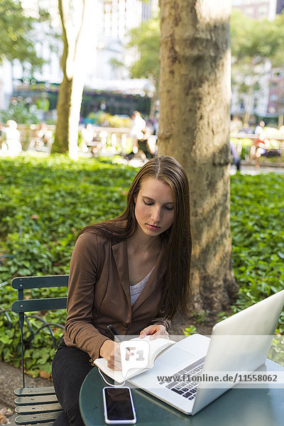 USA  New York  Manhattan  junge Geschäftsfrau mit Laptop am Tisch im Park und Notizen machend
