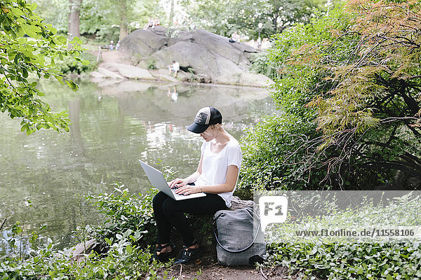 Junge Frau sitzt am Seeufer im Park mit Laptop