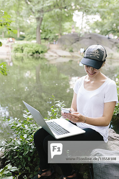 Junge Frau sitzt am Seeufer im Park mit Handy und Laptop