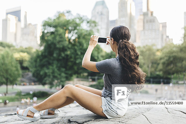 USA  Manhattan  young woman sitting in Central Park taking picture of skyline with smartphone