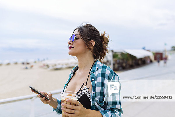 Young woman at the beach with cup of coffee and smart phone