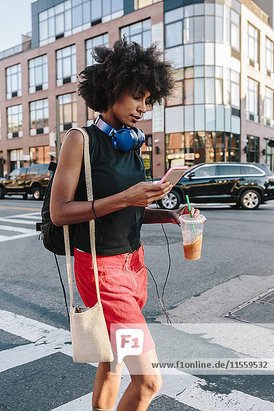 Young woman with headphones and smart phone crossong street in Brooklyn  carying take away drink