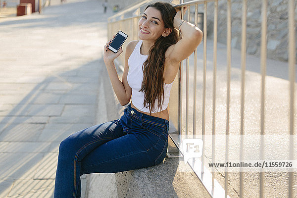 Portrait of smiling young woman with smartphone sitting on a wall
