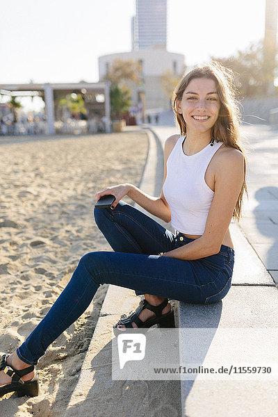 Spain  Barcelona  portrait of smiling young woman sitting on beach promenade at sunset